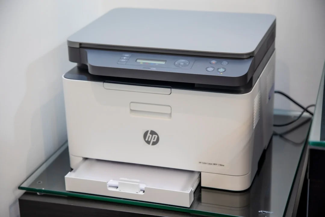 A high-volume office laser printer beside stacked OEM and compatible toner cartridges, with a cost-per-page comparison on a laptop screen in the background